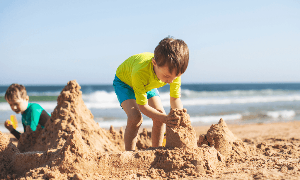 Two young children building sandcastles on a sunny beach, with a boy in a bright yellow shirt shaping a sandcastle in the foreground and another child playing in the sand beside the ocean.
