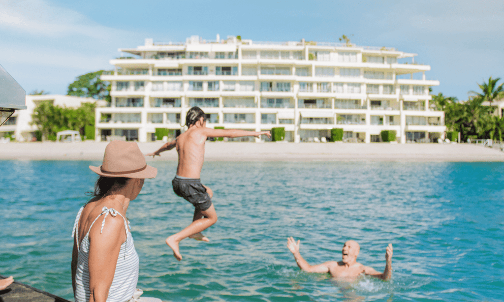 A boy leaps from a boat into the turquoise Noosa River toward a smiling man with outstretched arms, while a woman in a sun hat watches, with a white riverfront apartment building behind